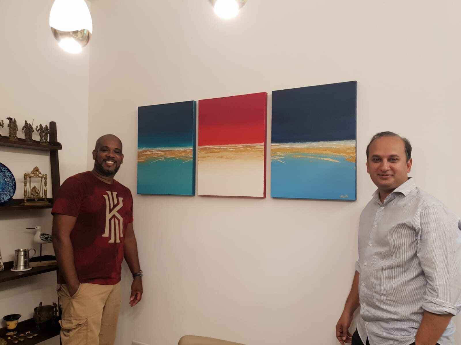 Raúl und ein Sammler neben einem Triptychon auf Auftrag – drei quadratische Meerbilder in Blau, Rot und Türkis mit goldenen Horizontlinien